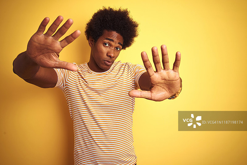 美国男子与非洲式头发穿条纹t恤站在孤立的黄色背景做框架用手手掌和手指，相机透视图片素材