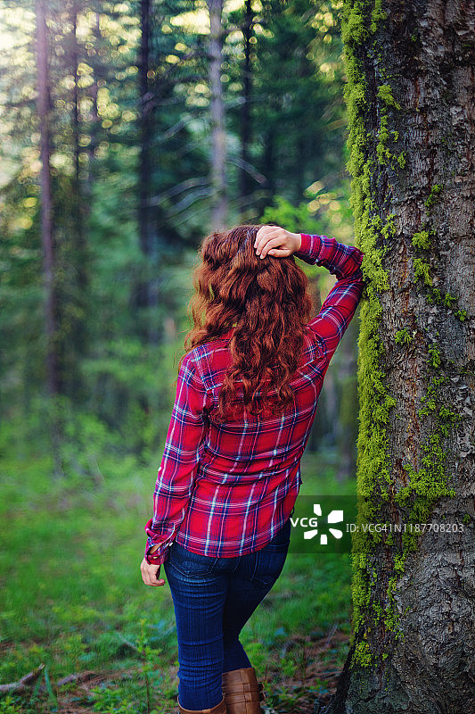 后视图的年轻女子在红色格子衬衫和牛仔裤倚在青苔覆盖的树在郁郁葱葱的森林图片素材