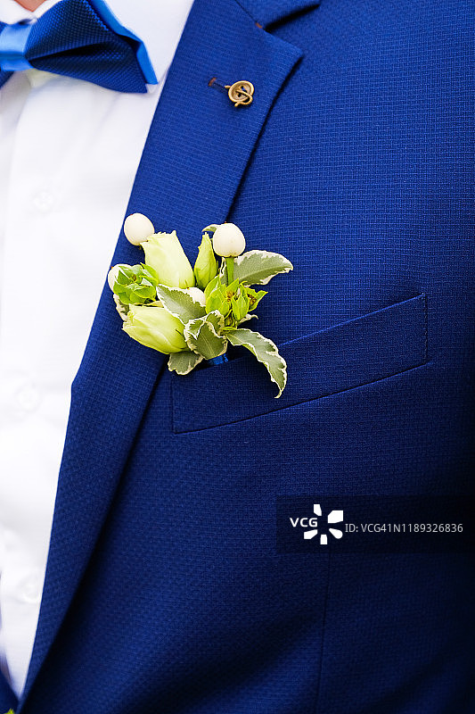 穿白色衬衫、领结和蓝色背心或夹克的年轻男子或新郎。背心口袋或翻领上有漂亮的白玫瑰和绿叶的胸花。婚礼的主题。图片素材