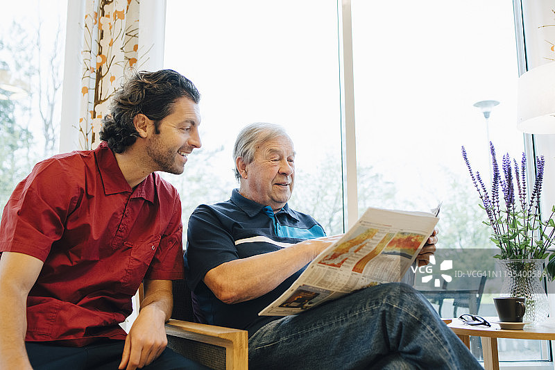 在退休之家，微笑的看护者坐在靠窗读报纸的老人旁边图片素材