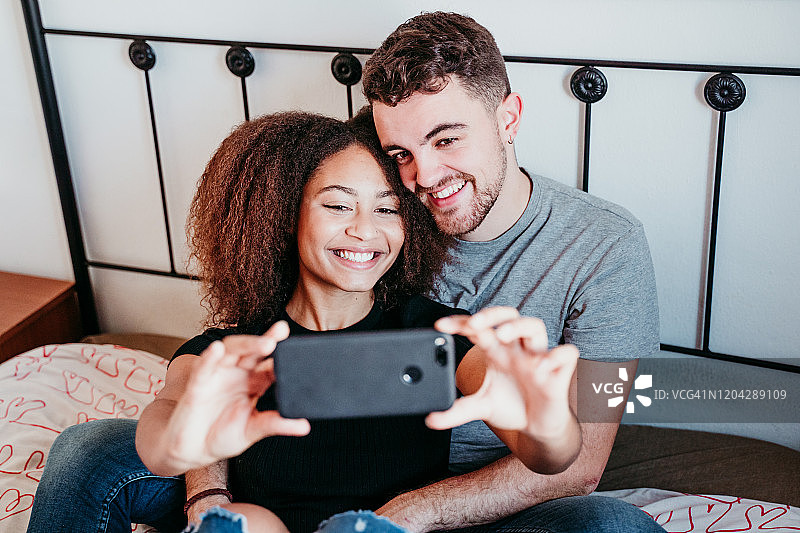 幸福的夫妻在家里相爱。美国黑人妇女和白人男子正在用手机自拍。爱民族概念图片素材