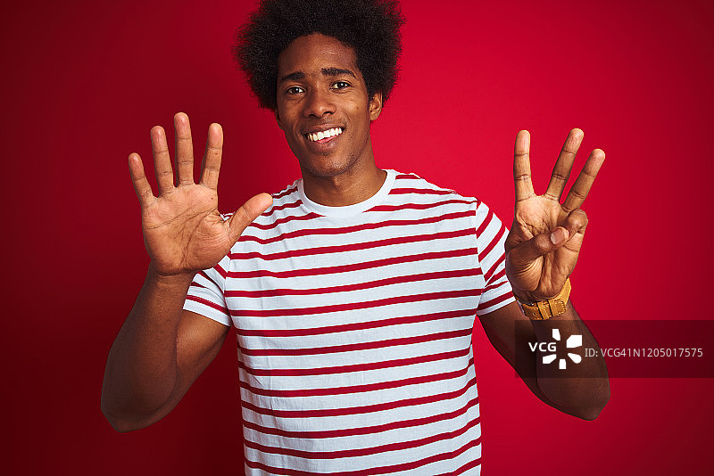 年轻的非洲裔美国人与非洲式头发穿条纹t恤在孤立的红色背景显示和指向第八位手指，同时微笑自信和快乐。图片素材