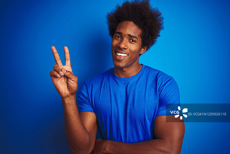 非洲裔美国人与非洲式头发穿t恤站在孤立的蓝色背景微笑与快乐的脸在镜头眨眼做胜利标志。二号人物。图片素材