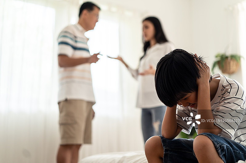 亚洲男孩坐在床上哭泣，而父母在家里打架或争吵冲突。孩子用手捂着耳朵不想听到暴力的争论。家庭中的家庭问题。图片素材