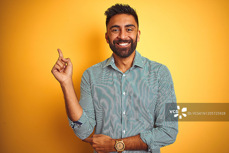 年轻的印度男子穿着绿色条纹衬衫站在孤立的黄色背景上，脸上带着大大的微笑，用手和手指指着一边看着摄像机。图片素材