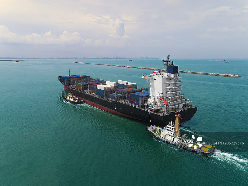 航拍俯视图拖船推着集装箱货船到码头在起重机桥上装卸货物，进行物流、进出口、航运或运输。图片素材