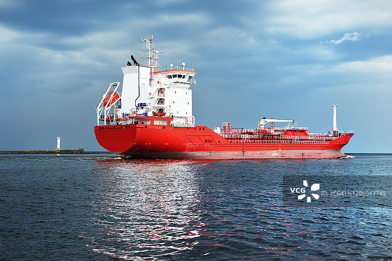 在开阔的波罗的海中装有起重机的大型红色货船。黑暗的乌云。背景是灯塔。拉脱维亚图片素材