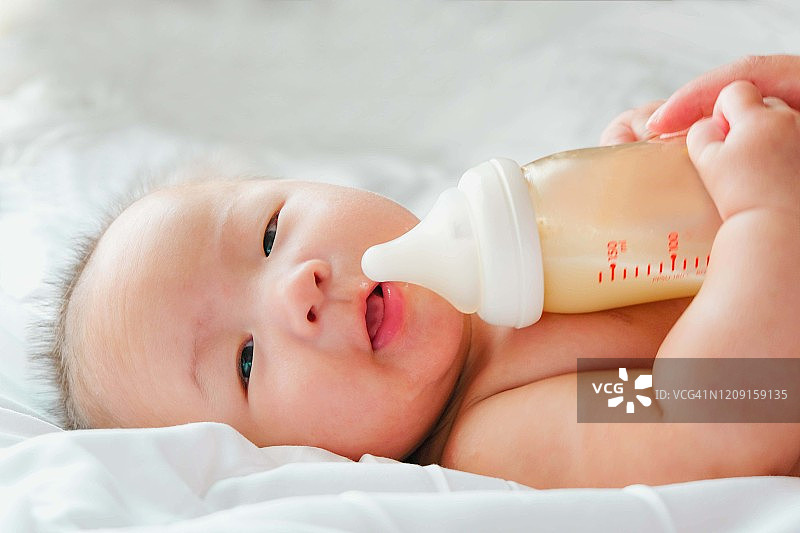 一个刚出生的亚洲婴儿在床上喝着奶瓶里的牛奶，可爱的黑眼睛婴儿躺在床上，一个孩子躺在床上休息图片素材