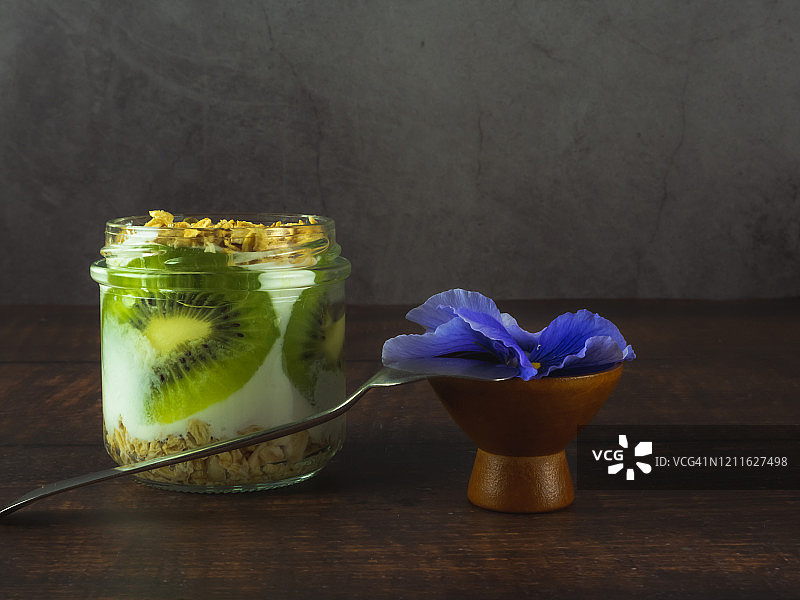 一杯含有猕猴桃和格兰诺拉麦片早餐的天然酸奶，在木桌上放一朵可食用的花和一把旧羊驼或银勺子图片素材