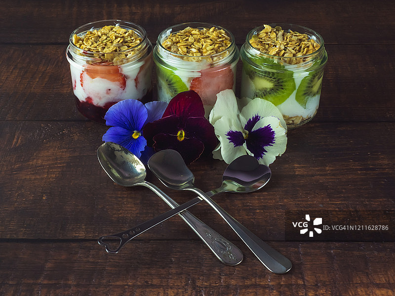 木桌上放着一杯天然酸奶、草莓、猕猴桃、格兰诺拉麦片早餐、可食用的鲜花和旧银勺图片素材
