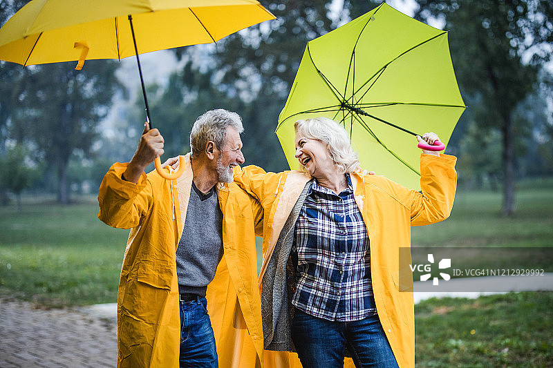 快乐的老夫妇穿着雨衣在公园里拿着雨伞玩。图片素材
