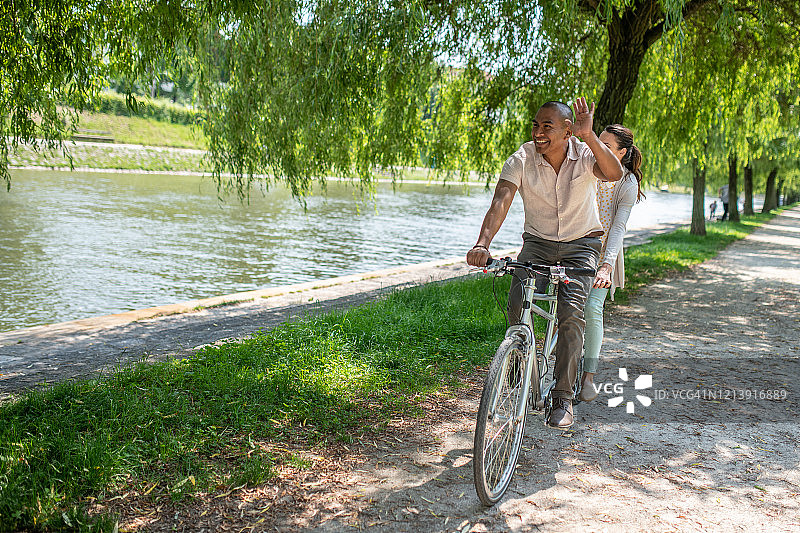 一对夫妇骑着双人自行车在河岸上挥手图片素材