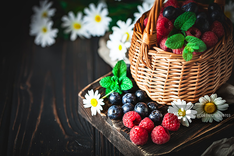 覆盆子和蓝莓装在一个篮子里，洋甘菊和叶子在黑暗的背景上。夏日与健康的饮食理念。背景与复制空间。有选择性的重点。图片素材