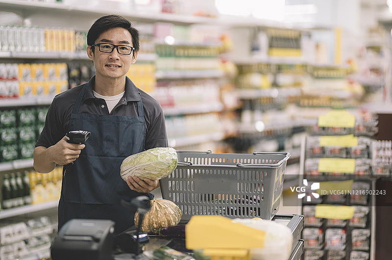 一位亚洲华人销售收银员拿着蔬菜、卷心菜和条形码阅读器站在收银台的收银台前，微笑着看着摄像头图片素材