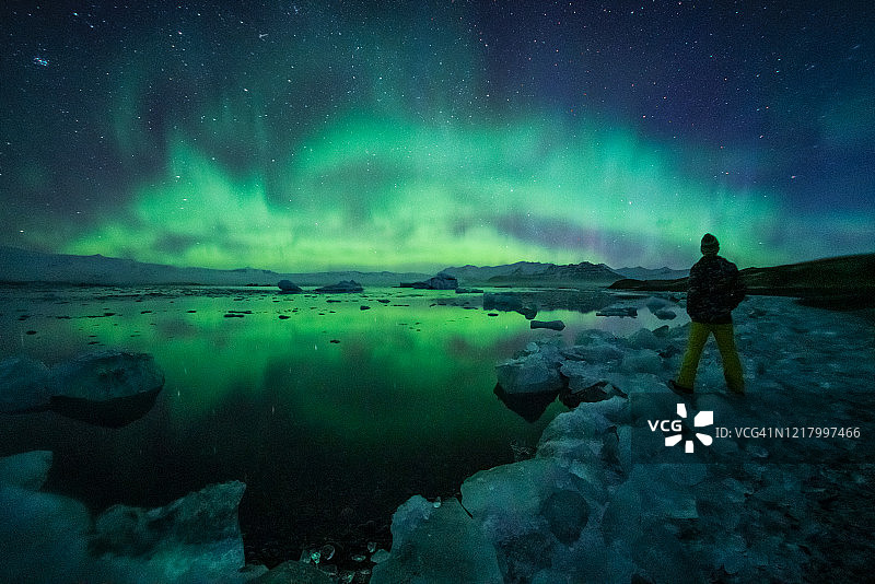 一名男子站在冰川泻湖上观看北极光，冰岛图片素材