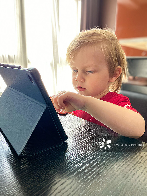 一个皮肤白皙的小孩坐在厨房的一张桌子旁，用平板电脑自学。图片素材