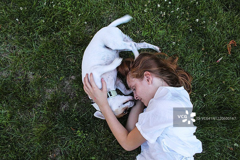 年轻的红发女孩和她的狗躺在草地上。他们都侧躺着，面对面。狗是白色的，她的衬衫也是白色的。这张照片是从她的腰部以上直接向下看的。图片素材