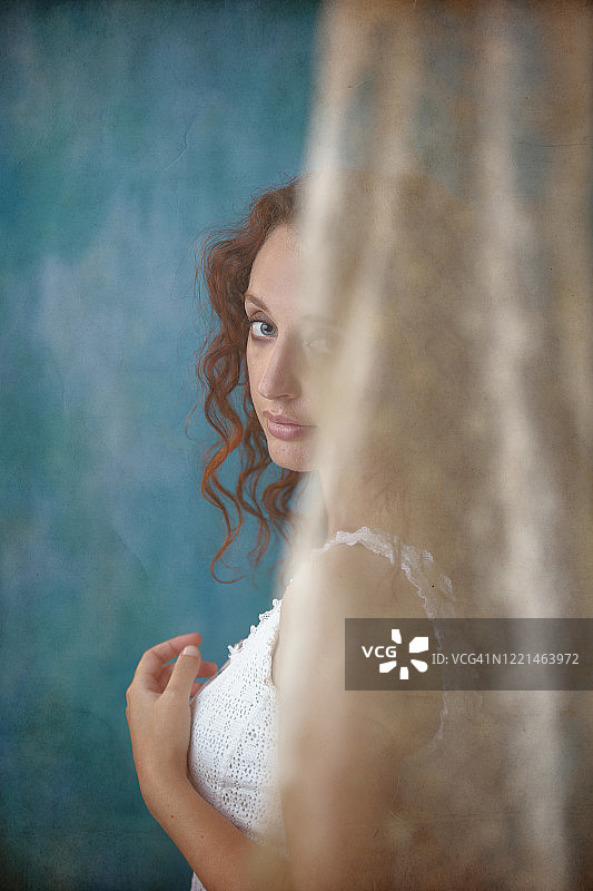 侧面的年轻女子波浪红头发穿着蕾丝无袖白色衬衫，半隐藏在花边窗帘后，看着相机，纹理的蓝绿色背景在工作室图片素材