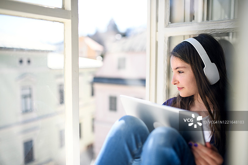 小女生戴着耳机和平板电脑，坐在家里的窗台上。图片素材