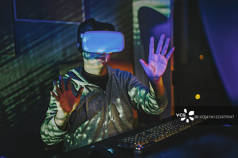 一名亚裔中国男性，晚上在他的家庭办公室自习室，戴上VR眼镜，在他的台式电脑前体验3D虚拟游戏图片素材