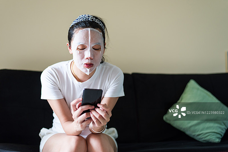 亚洲女性使用手机，同时在家里敷化妆面膜图片素材