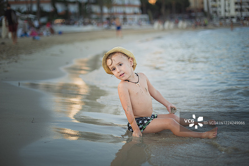 男孩坐在沙滩上，戴着帽子，看向别处图片素材