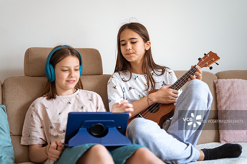 两个女孩在客厅里用着尤克里里琴、数码平板电脑和耳机图片素材