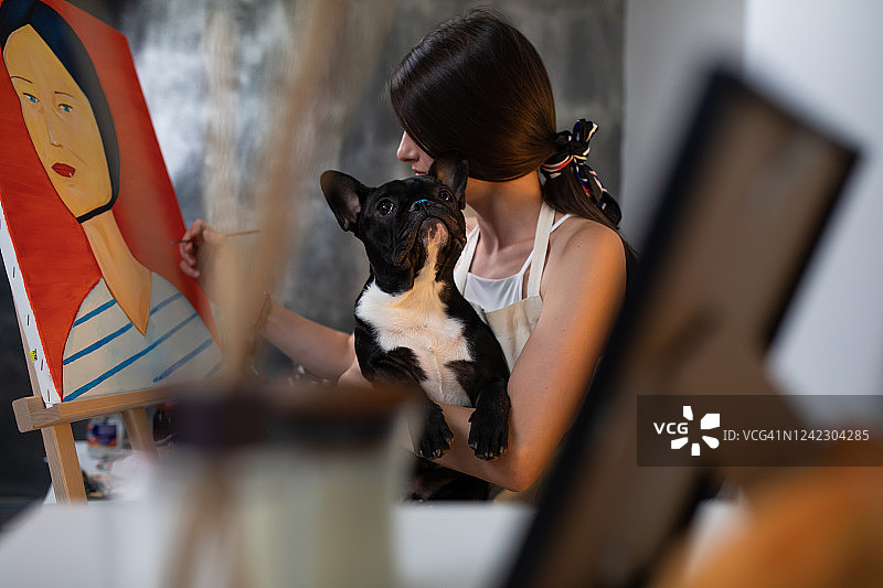 一只长着蓝色鼻子的可爱狗坐在一个年轻女子的腿上，而她正在画布上作画图片素材