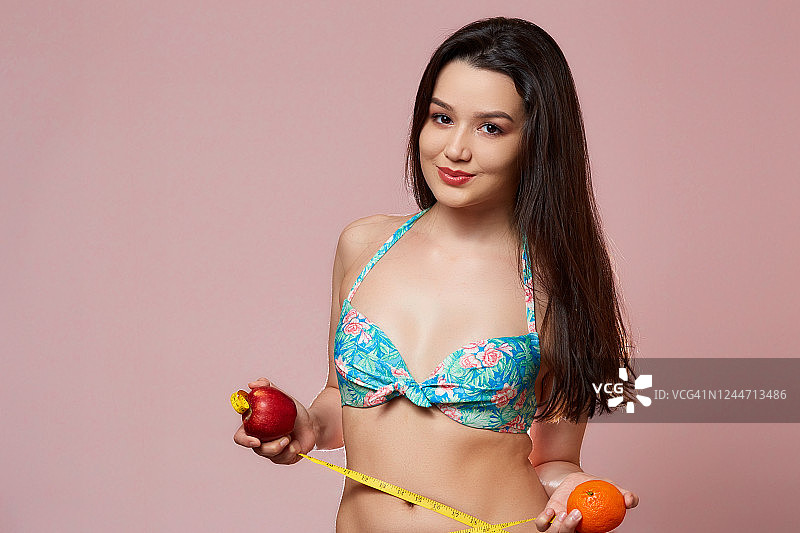 年轻美丽、苗条的亚洲女孩穿着泳衣。一只手拿着橘子，另一只手拿着苹果，手里拿着卷尺。在粉红色孤立的背景上图片素材