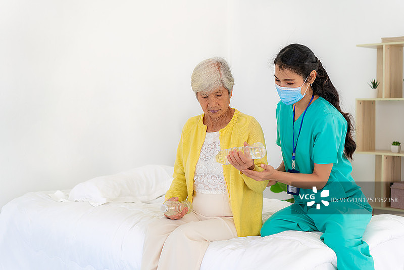 亚洲老年妇女关节炎在她的手腕接受关节炎物理治疗水瓶康复由专业护士或家庭健康助理在卧室在家里。图片素材