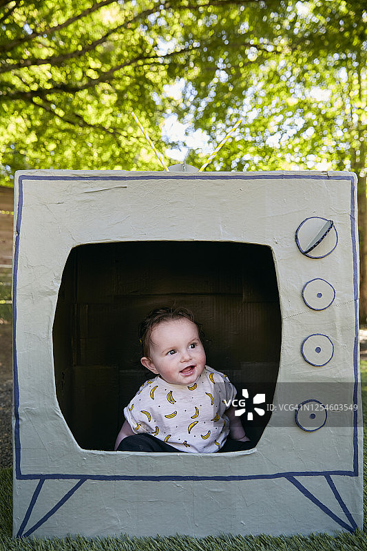 一个婴儿坐在硬纸板做的电视里，看着镜头微笑。图片素材
