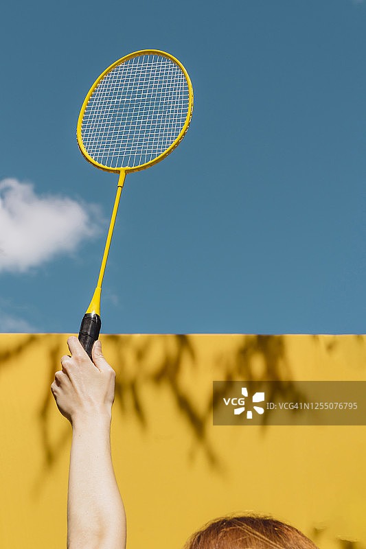 在阳光明媚的日子里，女子手持羽毛球拍越过黄色的墙壁，映衬着蓝色的天空图片素材