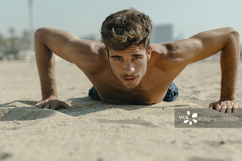 阳光明媚的日子里，一个赤膊的年轻人在沙滩上做俯卧撑图片素材