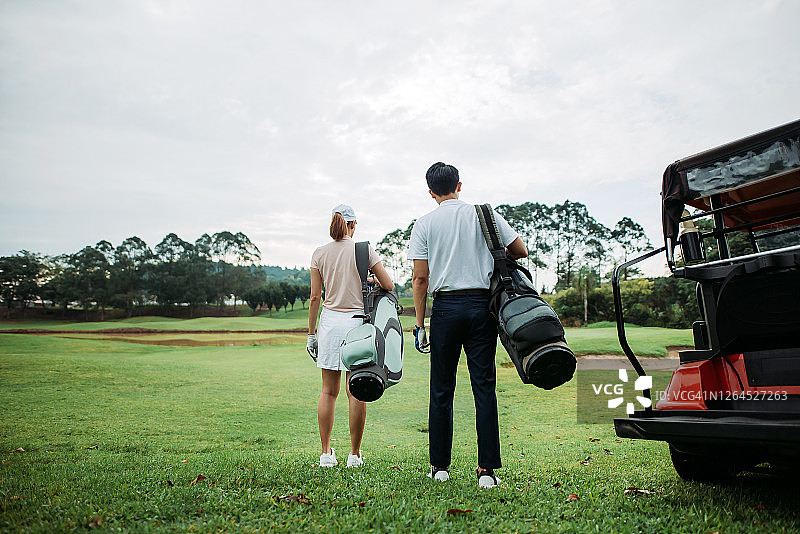 后视图的亚洲中国年轻夫妇高尔夫球手拿着高尔夫球袋，站在高尔夫球场的湖边看图片素材