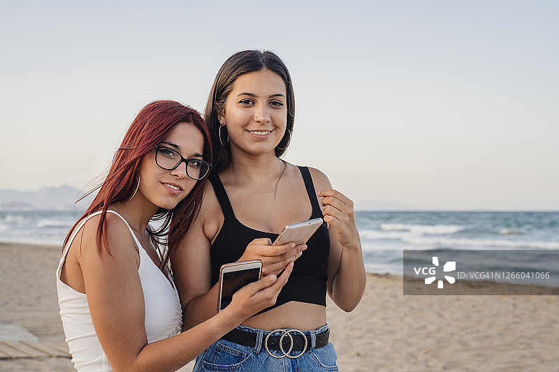 晴朗的天空下，十几岁的女孩在海滩上拿着智能手机图片素材