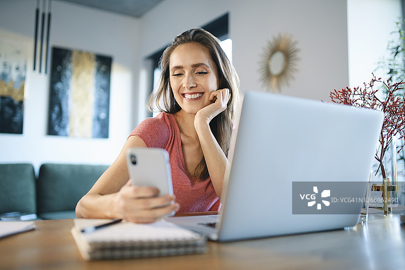 微笑的女性自由职业者使用智能手机的笔记本电脑在家庭办公室图片素材