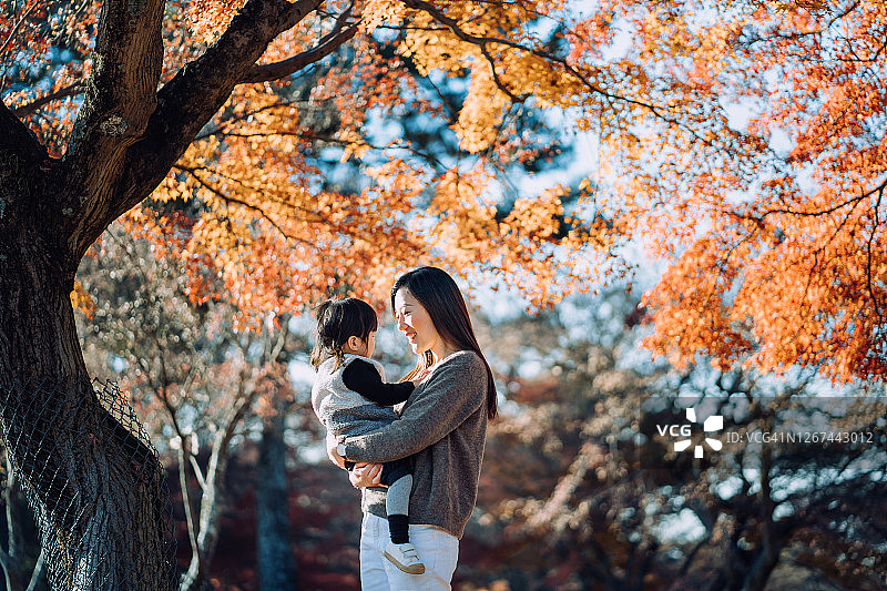 在一个阳光明媚的日子里，一位慈爱的年轻亚洲母亲抱着可爱的小女儿，一起在自然公园享受秋天的美景图片素材