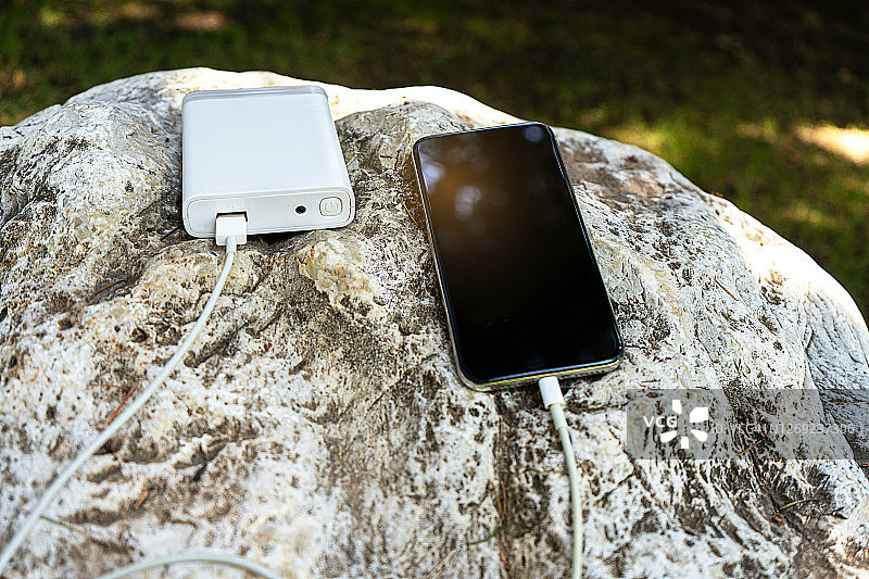 在一个阳光明媚的日子里，在岩石和绿草的背景下，通过设备共享电池无线充电的移动智能手机。图片素材