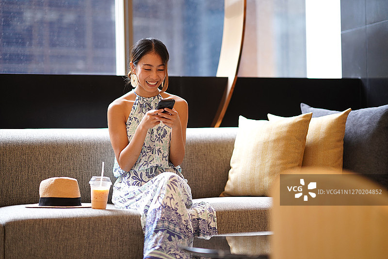 年轻的亚洲女性游客坐在沙发上与行李在酒店大堂使用智能手机聊天和微笑图片素材