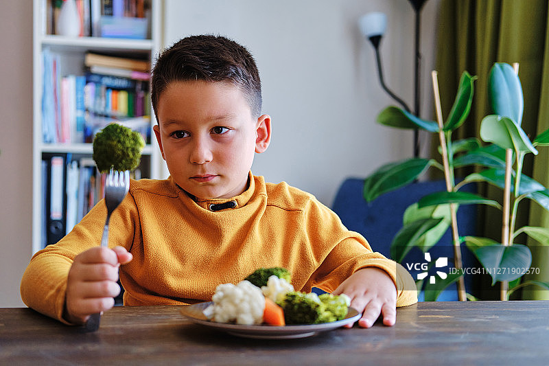 孩子对不得不吃蔬菜很不高兴。他的盘子里有许多蔬菜。他讨厌蔬菜。图片素材