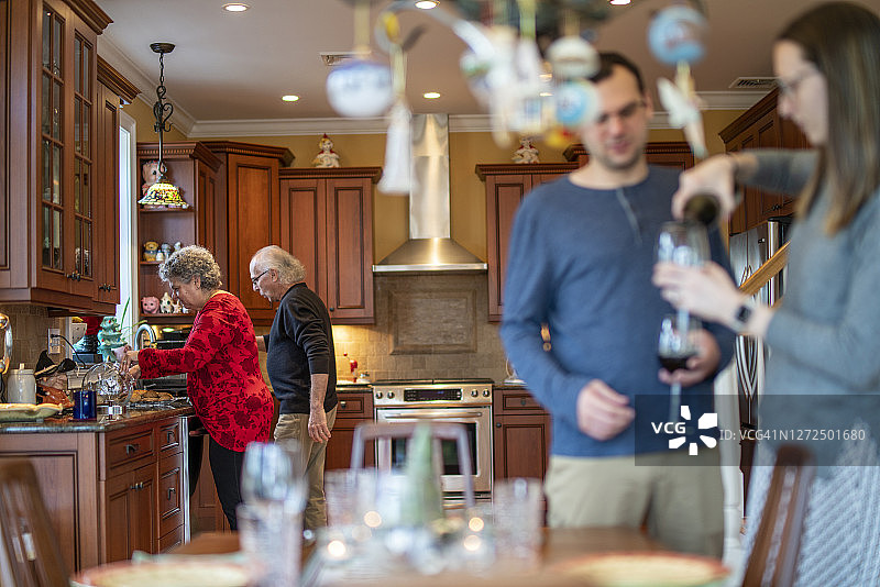 年轻女子为丈夫倒红酒，背景是年迈的父母在厨房工作图片素材