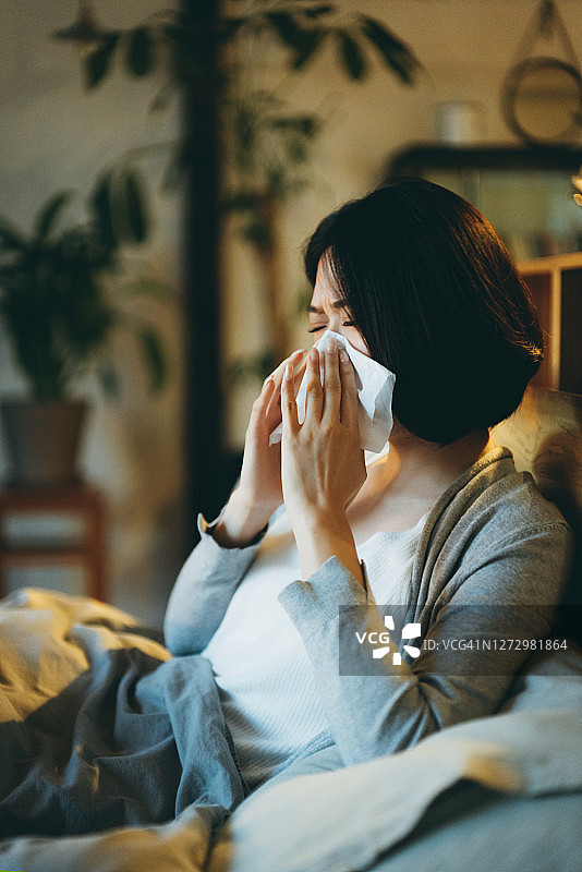年轻的亚洲妇女患感冒和流感，用纸巾擤她的鼻子，而躺在床上和在家休息图片素材