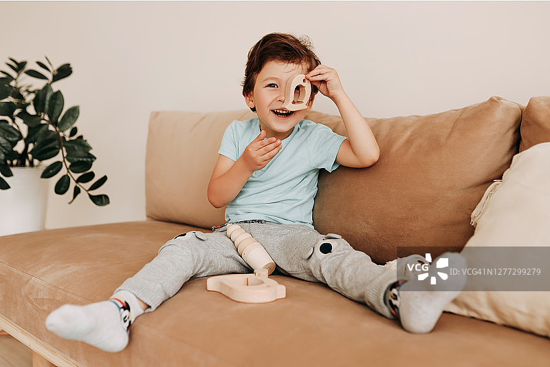 一个快乐的微笑的小男孩坐在舒适的家里的沙发上玩木制玩具，大笑图片素材