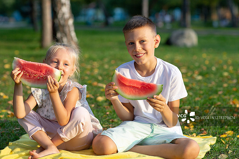 给可爱的孩子们，小男孩和小女孩在秋天的公园草地上吃着多汁的西瓜图片素材