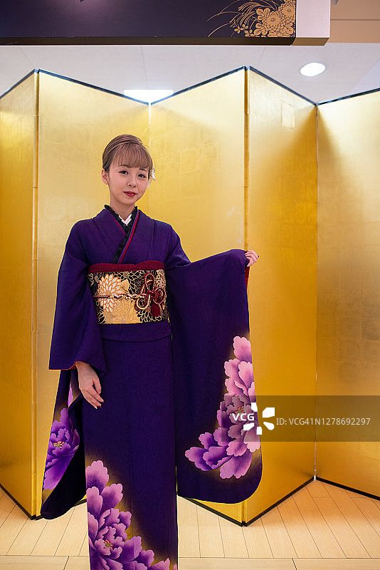 身着Furisode和服的日本妇女站在“Seijin Shiki”成人礼的金色“Byo-bu”折叠屏风前的肖像图片素材