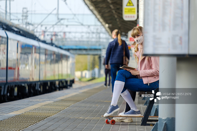 一个戴着保护面罩的金发少女坐在火车站的长凳上看智能手机。图片素材
