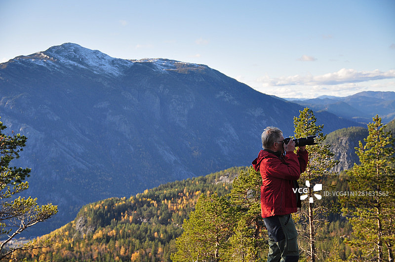 穿着红色夹克的老人在山上拍照。山顶上洒满了新雪。后面是山脉。图片素材