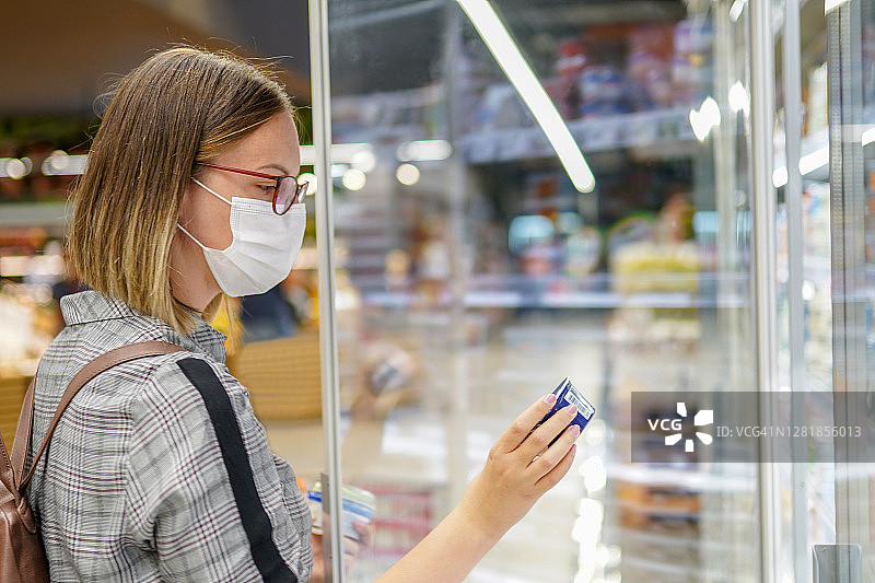 戴着面罩和眼镜的女人站在超市的冰箱前阅读营养标签图片素材