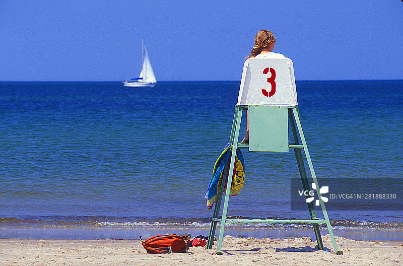 海滩上的女救生员坐在高高的救生员椅上看着一艘白色的帆船驶过图片素材