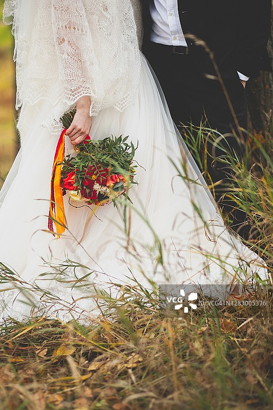 新娘手持一束美丽的玫瑰，有白的，有黄的，有红的，还有绿叶。新婚夫妇拥抱，新郎和新娘的手特写，背景是干枯的枯草。秋季婚礼的主题。图片素材
